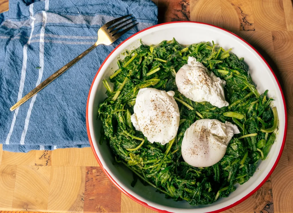 White plate with wilted greens and poached eggs, set on a wooden table with a blue napkin nearby and a fork overhead.