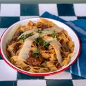 Bowl of pasta with wide ribbon noodles, meat sauce, shaved parmesan, and herbs on a checkered table.
